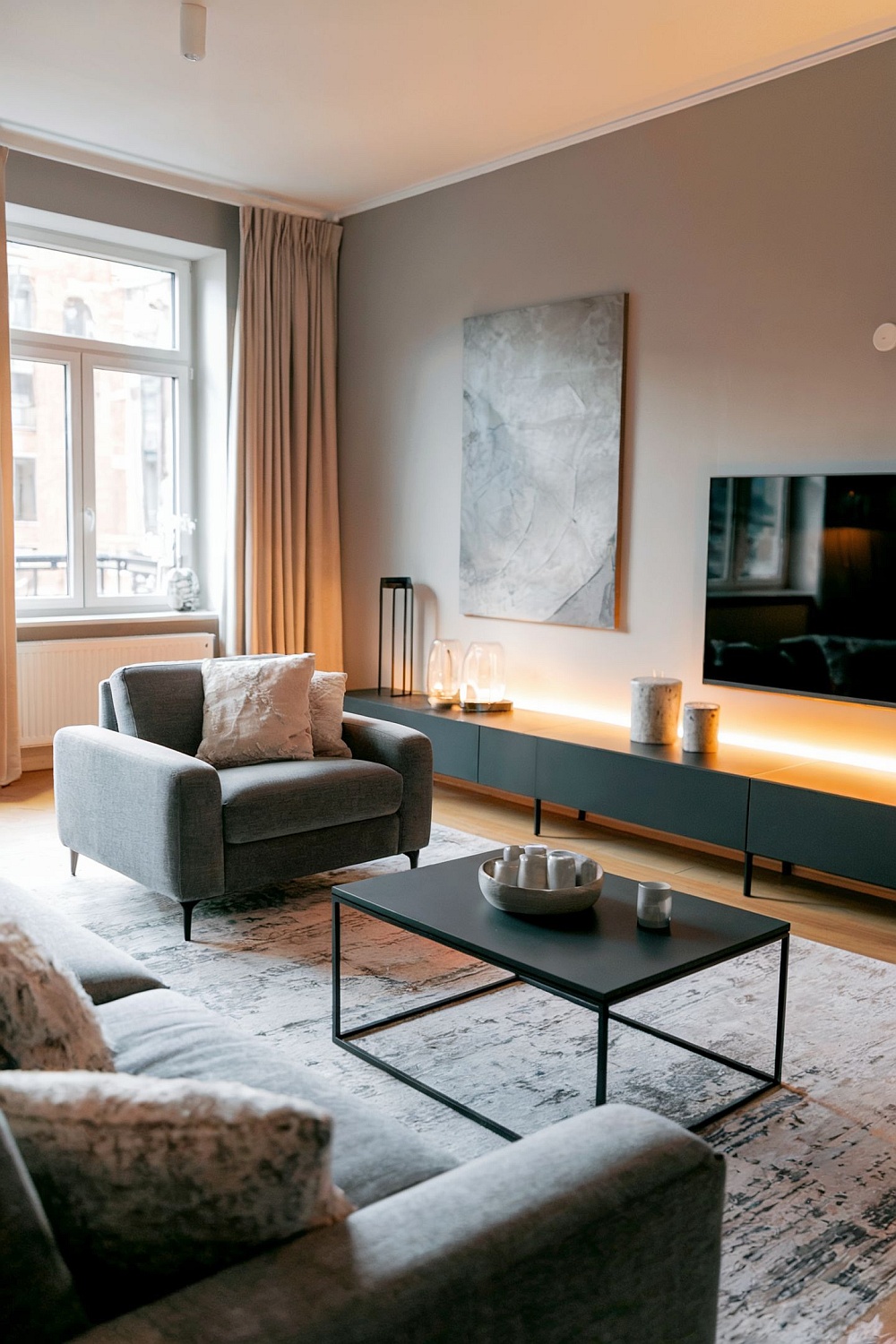 Stilvolle graue Wohnzimmer Ideen mit neutraler Wohnzimmergestaltung, gemütlichem grauem Sessel und Sofa, indirekter moderner LED-Beleuchtung an der TV-Wand, minimalistischem Couchtisch und eleganten Accessoires sowie Wanddekorationen.