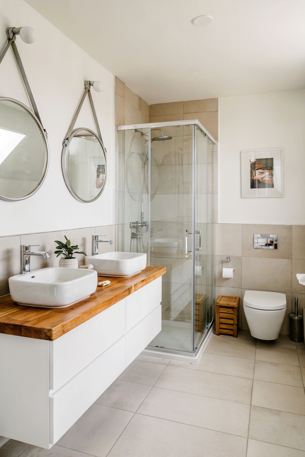 Modernes Badezimmer mit Doppelwaschbecken auf Holzplatte, Glasdusche, Wand-WC und neutralen Fliesen in Beige und Weiß.