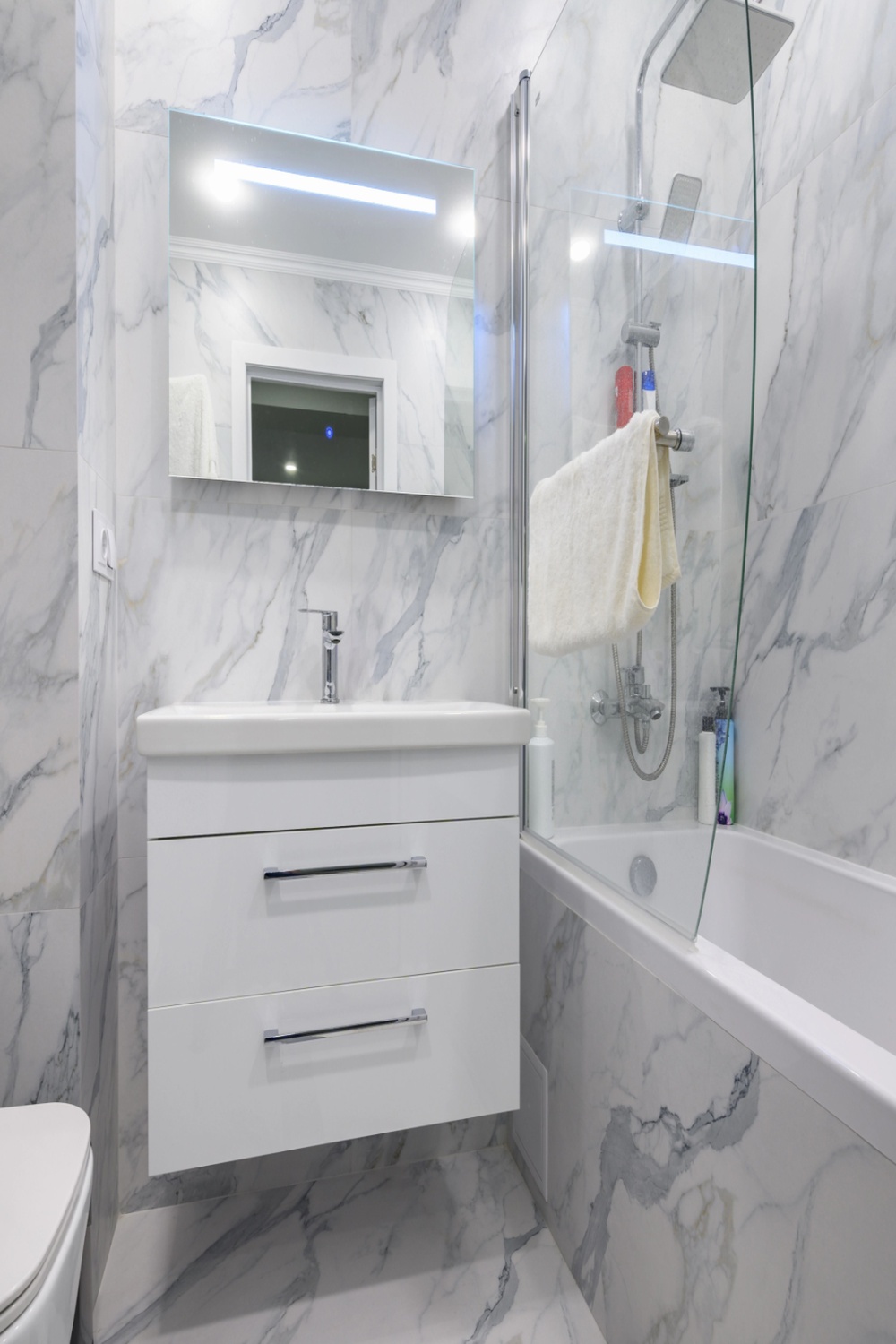 Kleines Badezimmer mit weißen Marmorfliesen, modernem weißen Waschtisch, Spiegel und Badewanne mit Glaswand.