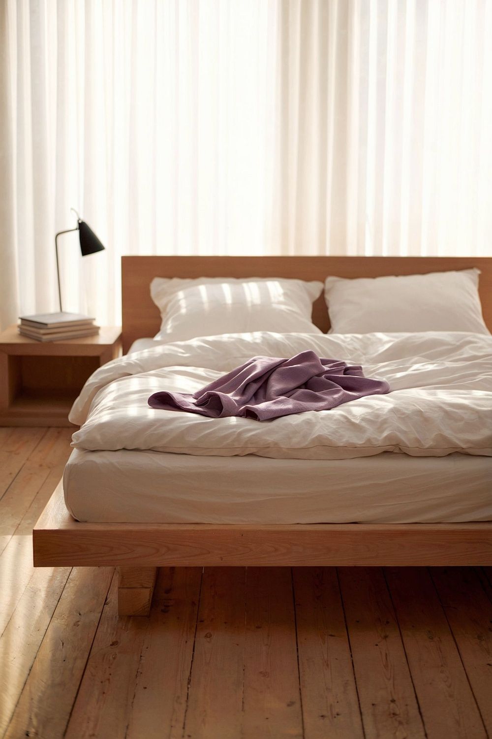 Ein minimalistisches Schlafzimmer mit einem Bett aus hellem Holz, weißer Bettwäsche und einer violetten Decke. Die warmen Holzböden und das natürliche Licht durch die Vorhänge schaffen eine beruhigende Atmosphäre.