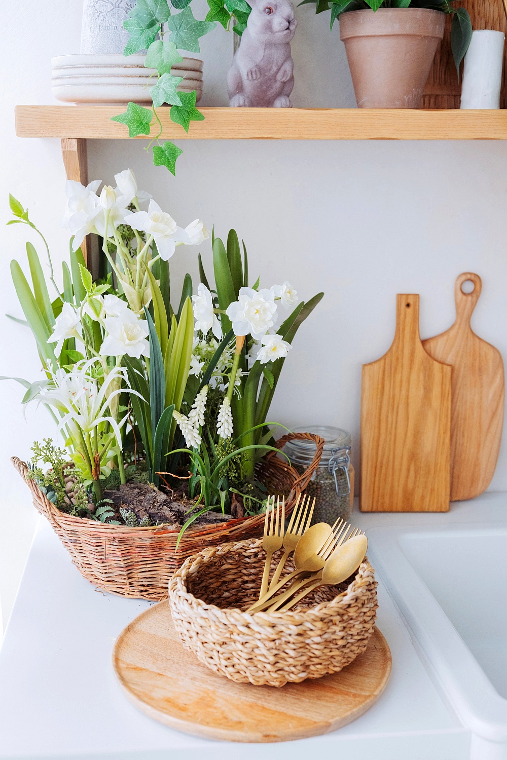Ein geflochtener Korb mit weißen Blumen und grünen Blättern, daneben rustikale Holz-Accessoires wie Schneidebretter und Körbchen.