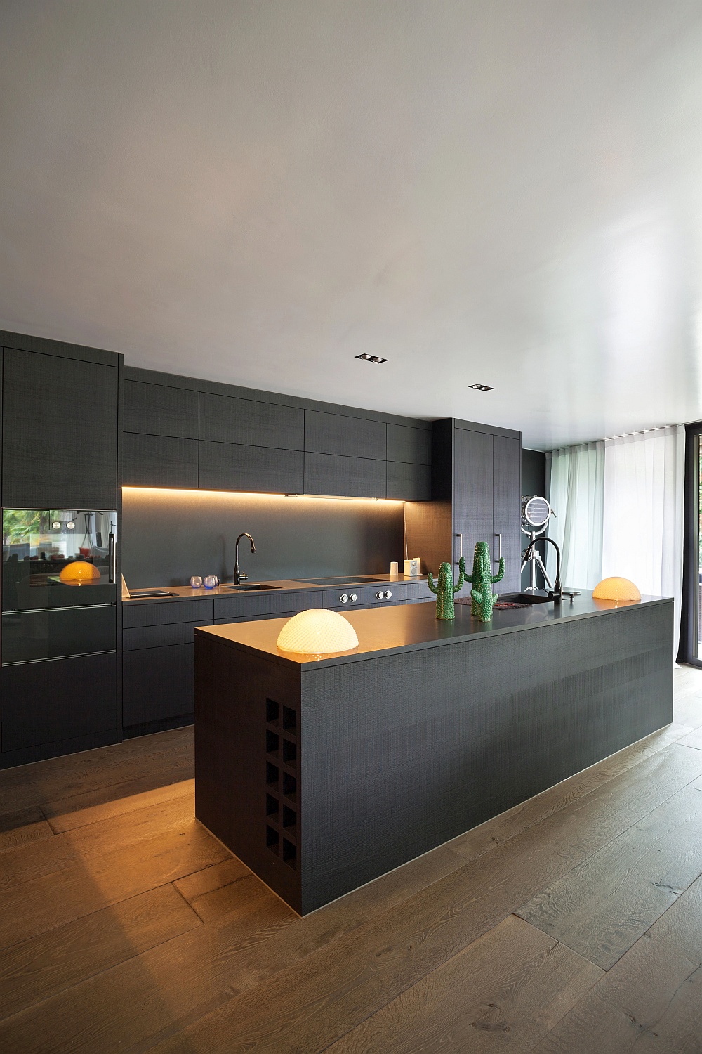 Eine luxuriöse, moderne dunkle Küche mit viel dunkelgrau und anthrazit, grifflosen Schränken, integrierten Geräten, einer langen Kücheninsel, minimalistischen Lampen und einem hellen Holzboden.