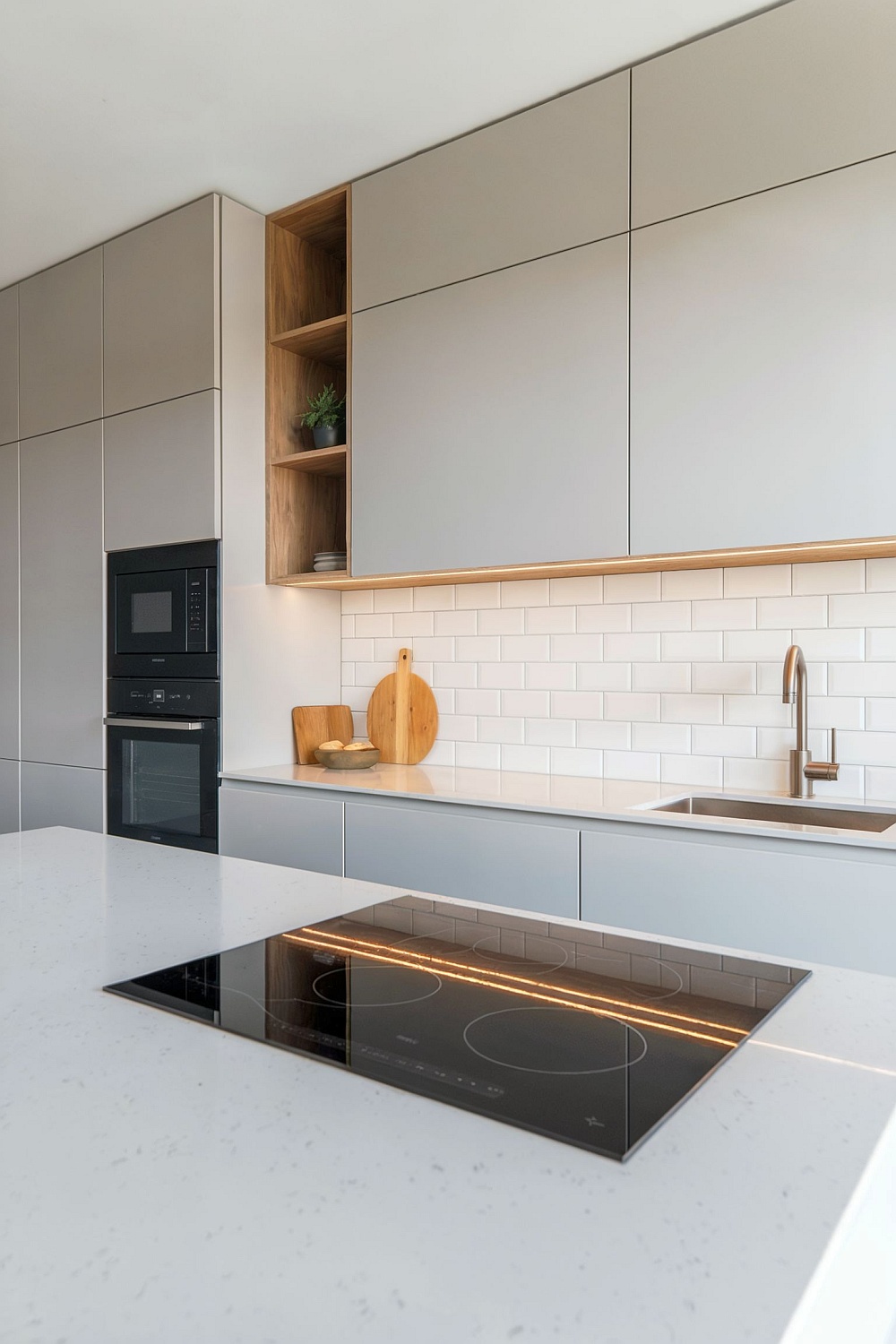Eine moderne Küche in hellgrau mit grifflosen Fronten, integrierten Holzregalen, einer weißen Metrofliesen-Rückwand, und einer Kücheninsel mit Kochfeld.