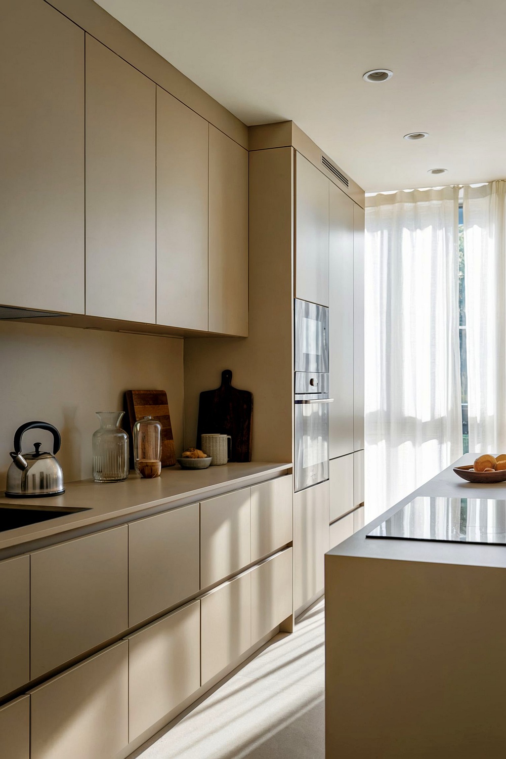 Eine minimalistische Küche in mattem Sandgrau mit grifflosen Schränken, integrierten Geräten, einer schlichten Arbeitsplatte und einem Fenster mit Vorhängen.