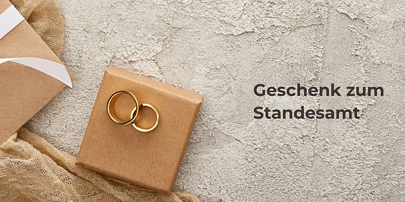 Geschenk zum Standesamt: zwei goldene Eheringe auf einer Geschenkschachtel neben einem Umschlag mit weißer Schleife