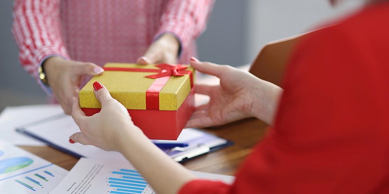 Weihnachtsgeschenke für Kollegen: Kollegin überreicht ein goldenes Geschenk mit roter Schleife am Schreibtisch