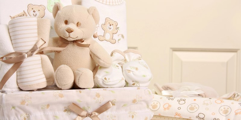 Geschenk zur Babyparty: Korb mit Teddybär, Babykleidung und Schühchen auf hellem Bettzeug