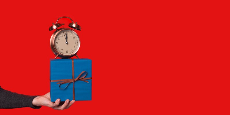 Last Minute Geschenk: Hand hält ein blau verpacktes Geschenk mit Wecker darauf vor rotem Grund